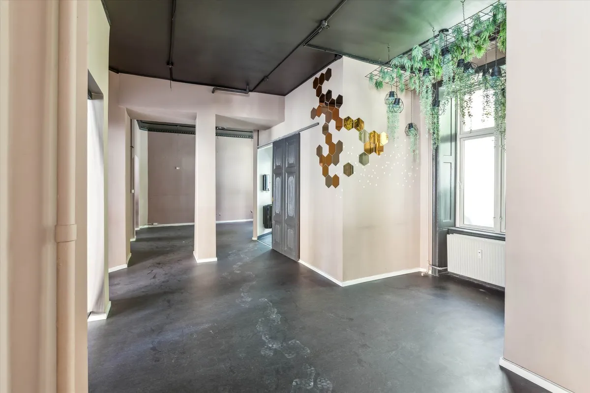 En tom gangvej med højt loft, gulvet er gul og lyse hjørner. Der er to døre og et gulv med en plante på væggen. Der er et