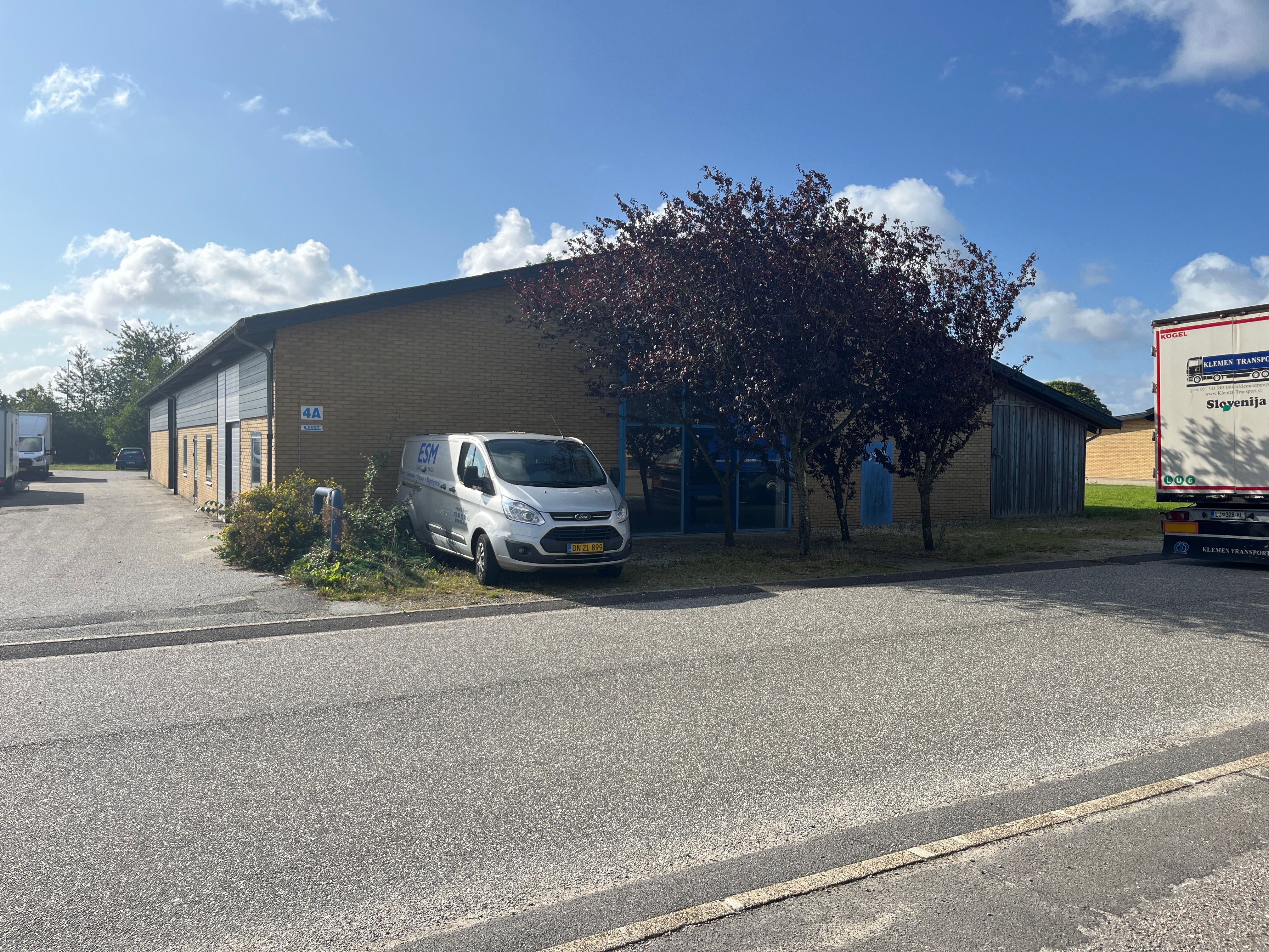 Lejemål i Stilling med parkeringsplads og skur, med to køretøjer parkeret og en stor skov med mørke løv og lyserøde blade.