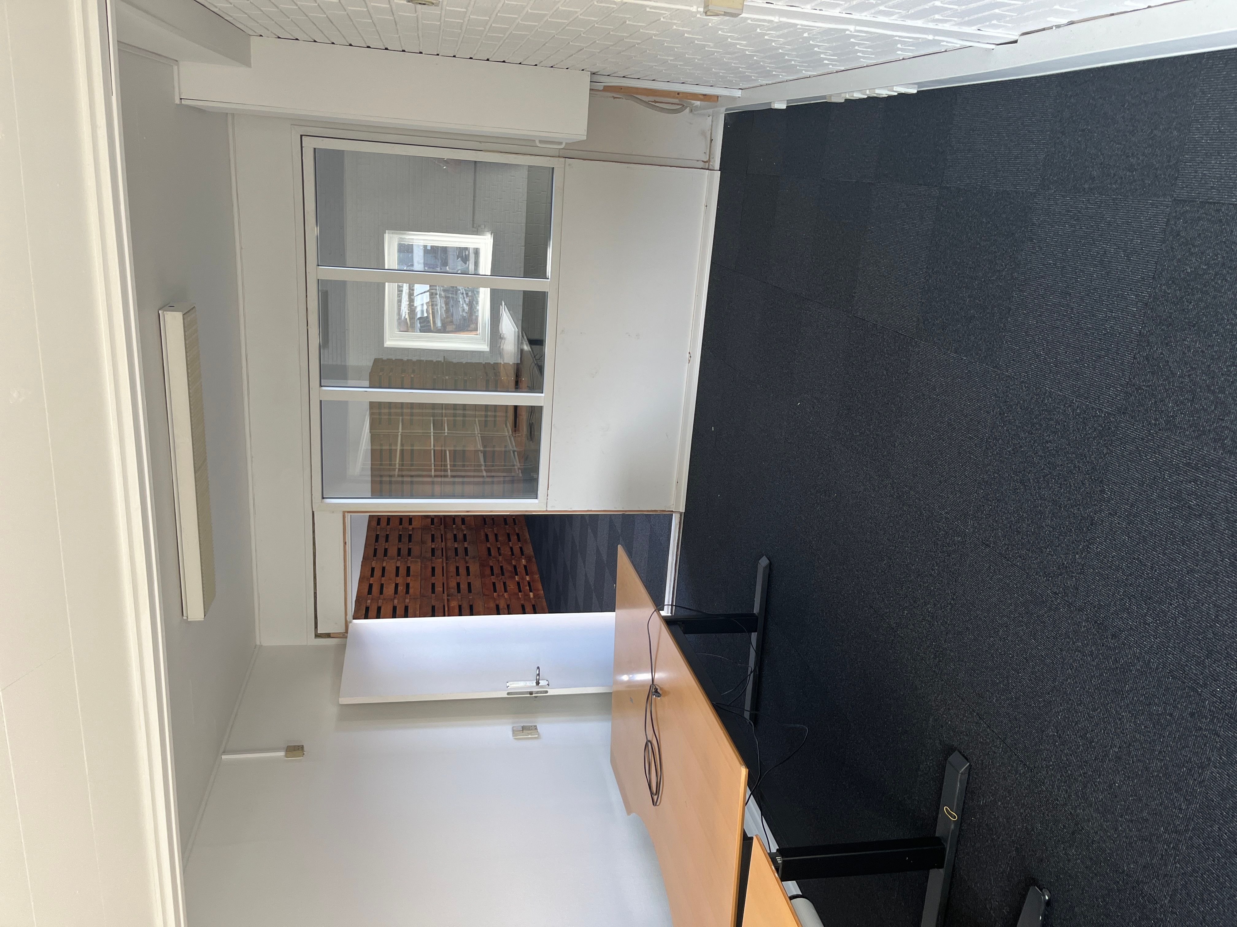 Billede af et tomt kontor med trægulve, hvide vægge og mørkt gulvtæppe. Værelset er ubenyttet og har glasdøre og vinduer.