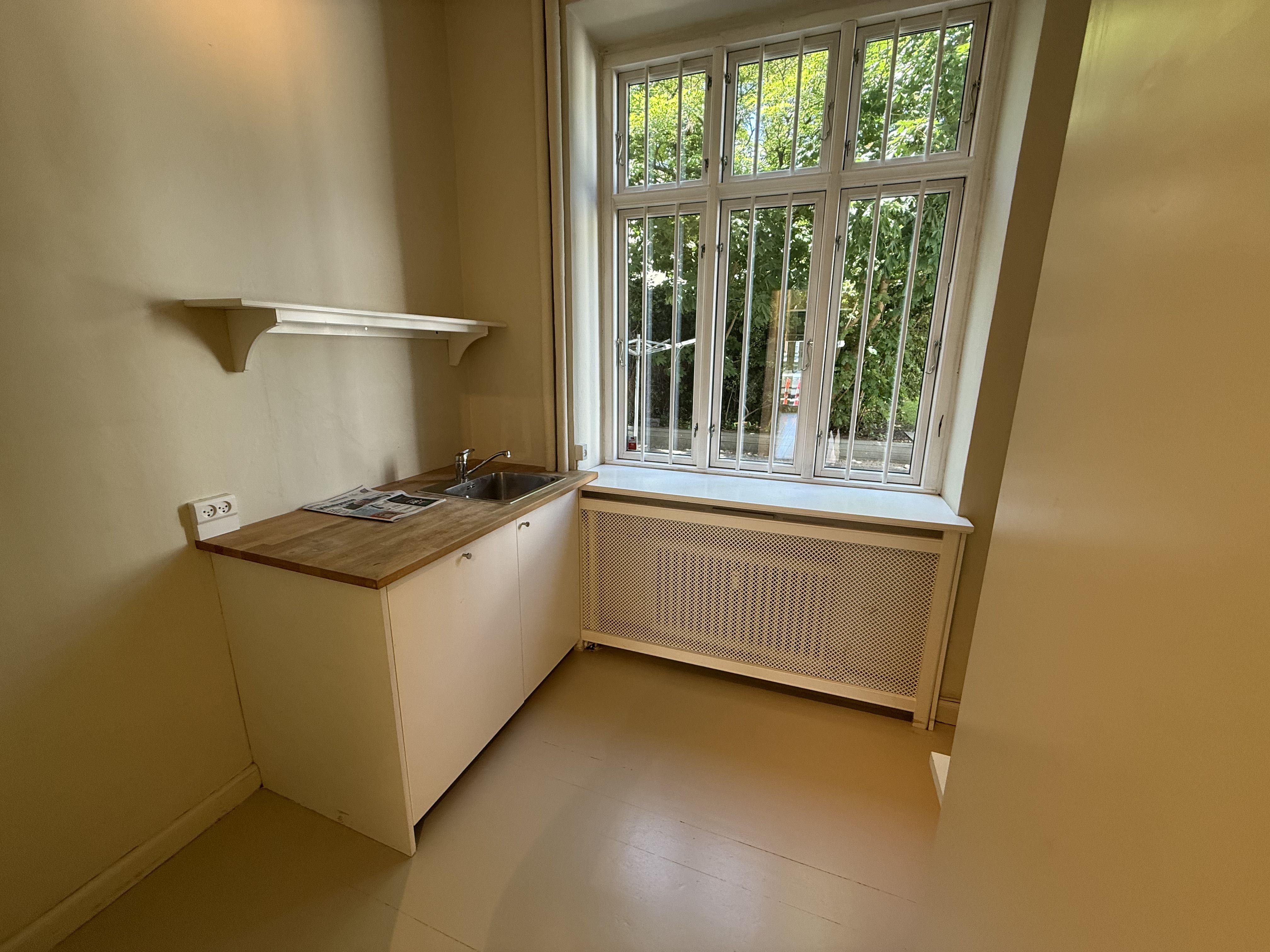 Et lille køkken med hvidt køkkenskab, brædt på bordpladen, trægulv, radiator og et vindue med barstange og udsigt til grønne