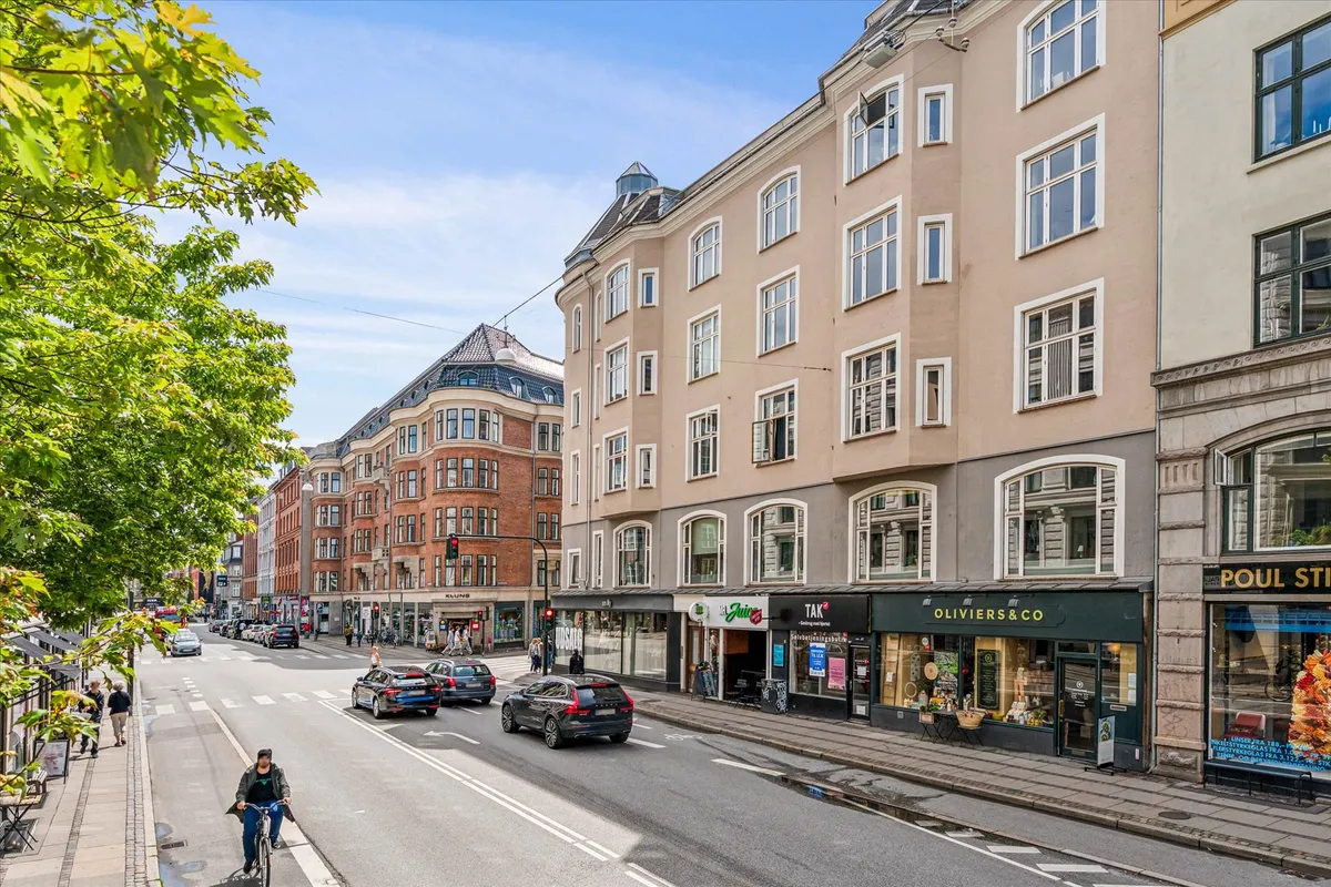 Mennesker går forbi butikker på en bred gade med biler og cykelryttere i en by