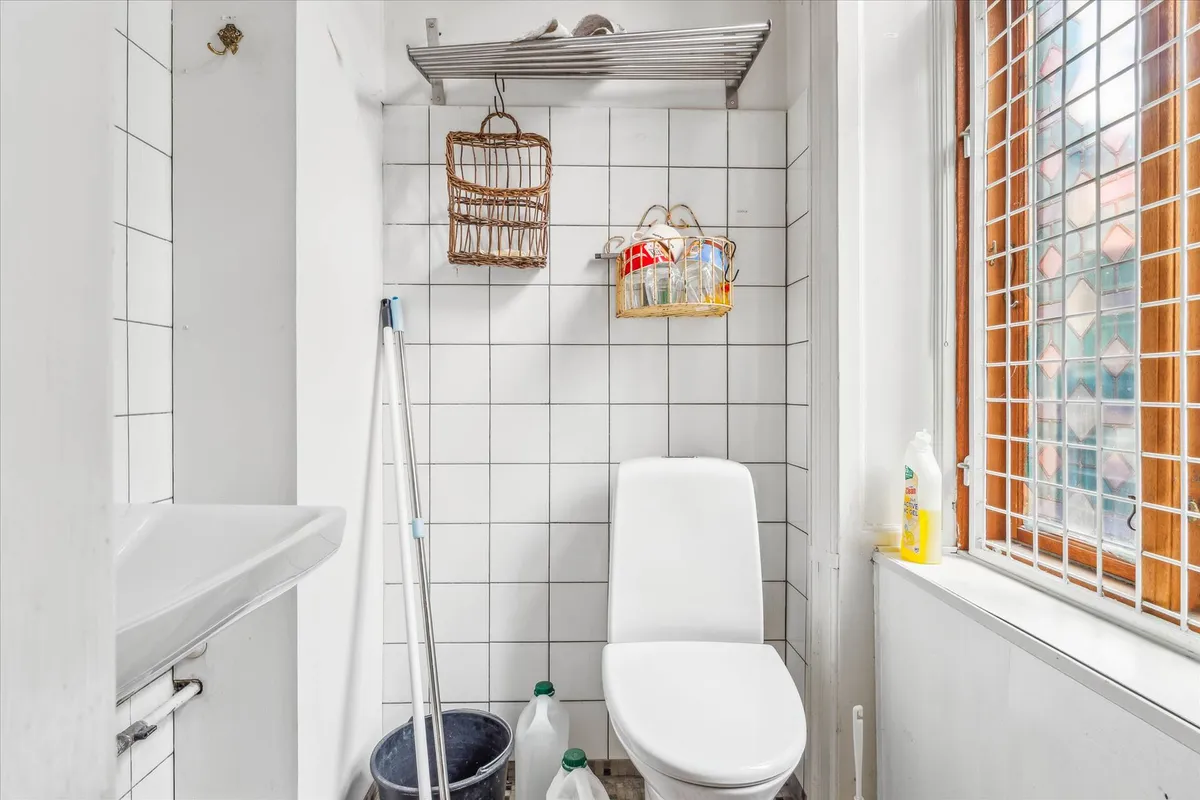 Et lille badeværelse med hvide kakeloverflader og en toilet, et rengøringssæt og to kurve hængende på væggen