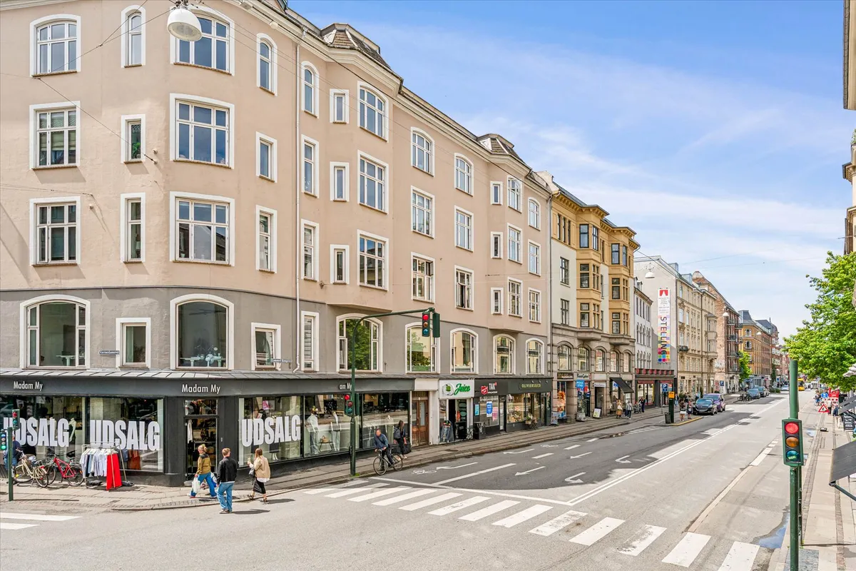 En bred gågade med forretninger, hvor folk går og cykler. En rødmaling og gultrafiklys er synlige. Et træ og nogle buske