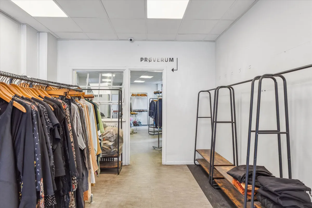 Genbrugsbutik med tøj og tøjstandere. Sort og hvid væg med Proverrum-tekst. Indgang med tøj på hængere og tøjstandere. Tøj