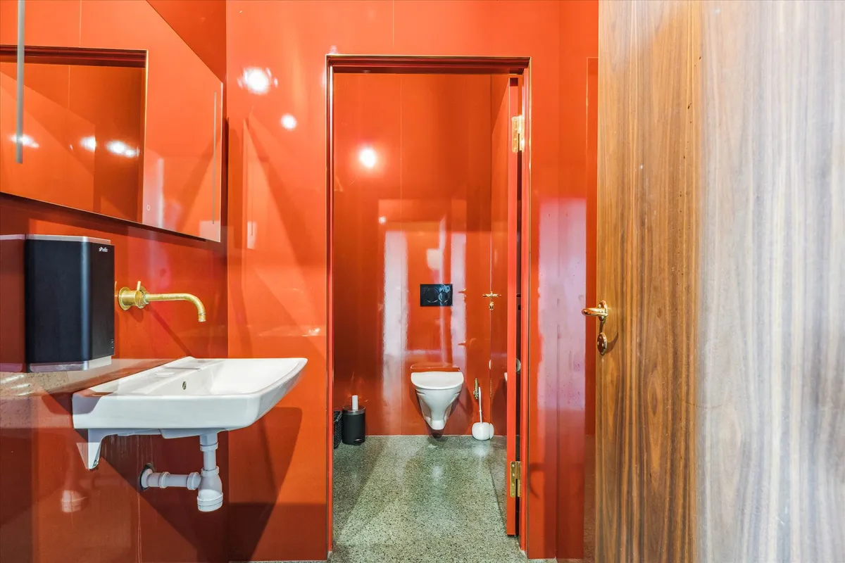 Toiletrum med orange vægge, hvidt toilet, hvidt vask, guldfarvet vandhane og glasdør