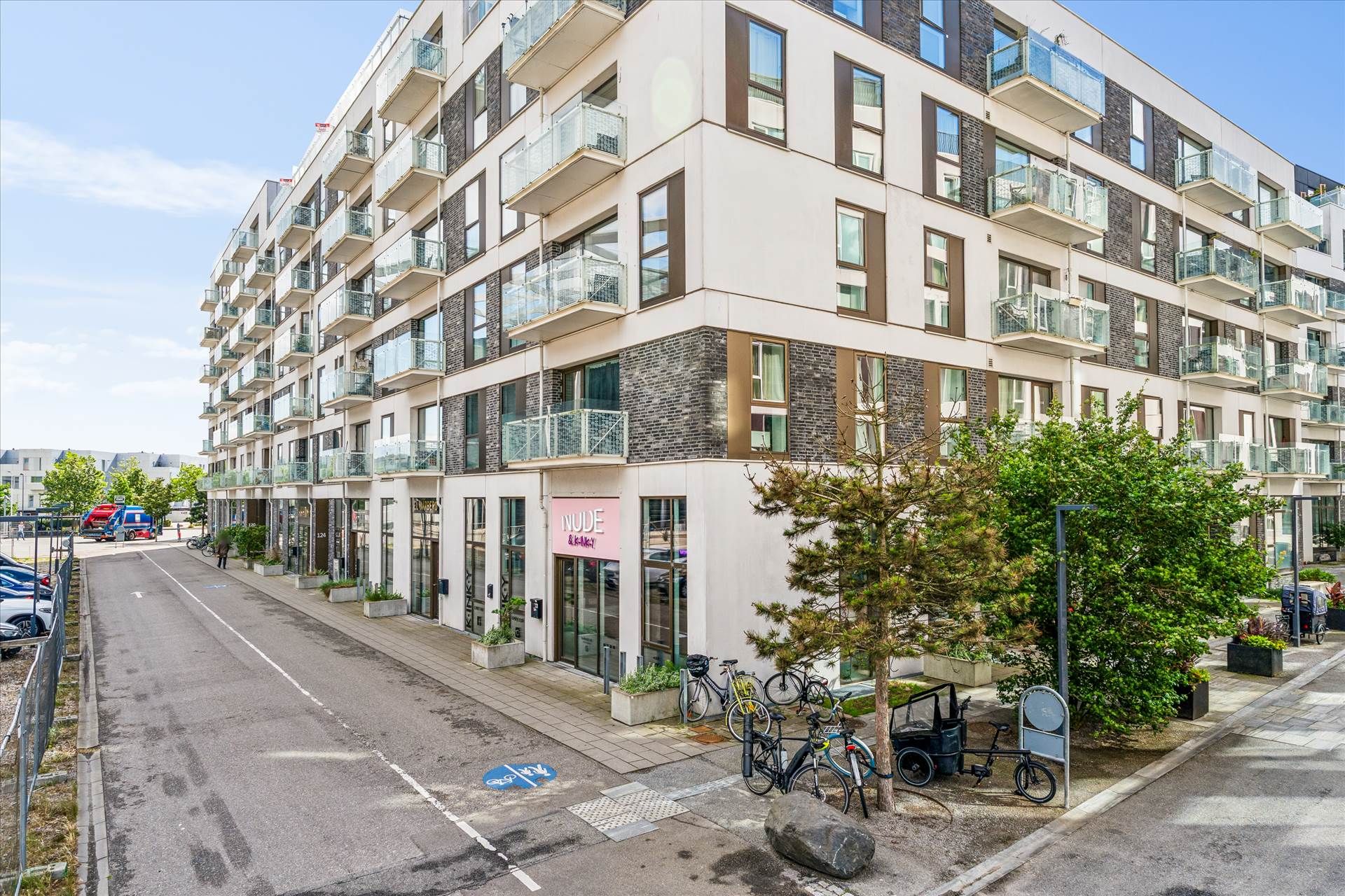 Butik med skriften Nude & Gay på facaden. Lejemål med parkerede cykler og grønne planter. Bygning med terrasser og