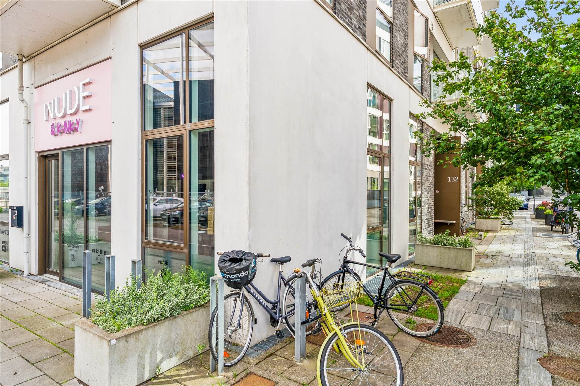 Et butiksvindue med en pink farve og en tekst, der læser "Nude & Kinky" og nogle planter og cykler foran butikken