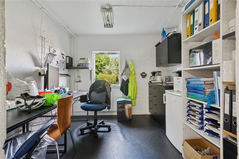 Et kontorlokal med skrivebord, stol, computer, mikroovn, hylder med mapper og skriftlig kommunikation samt en vinduesåbning