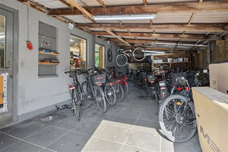 En række cykler er parkeret i en garage. Cyklerne er i forskellige farver og størrelser. Garagen har træloft og en stor