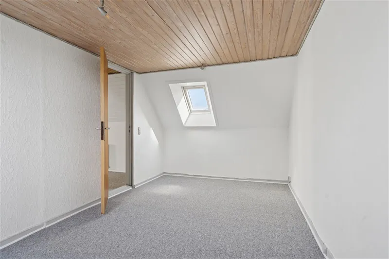 En tom værelse med lysegrå gulvbelægning og hvide vægge samt loftsvindue og åben dør til et andet værelse