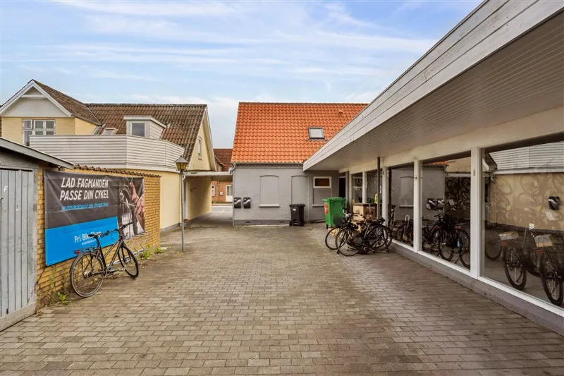 En bred gadegade med fire huse, et rødt tag og en plakat med teksten Lad Fagmanden Passe Din Cykel. En række cykler står