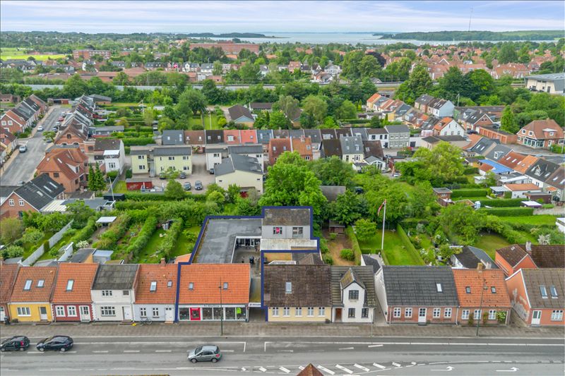 Byggegrund med handelslokaler i Middelfart. Bygninger med mange vinduer og garager i baghaven. Biler kører på vejen foran.