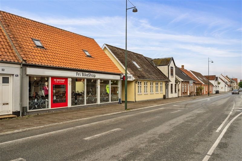 En bred tosporet gade med en Fri BikeShop og andre butikker i Middelfart, med solnedgang på himlen