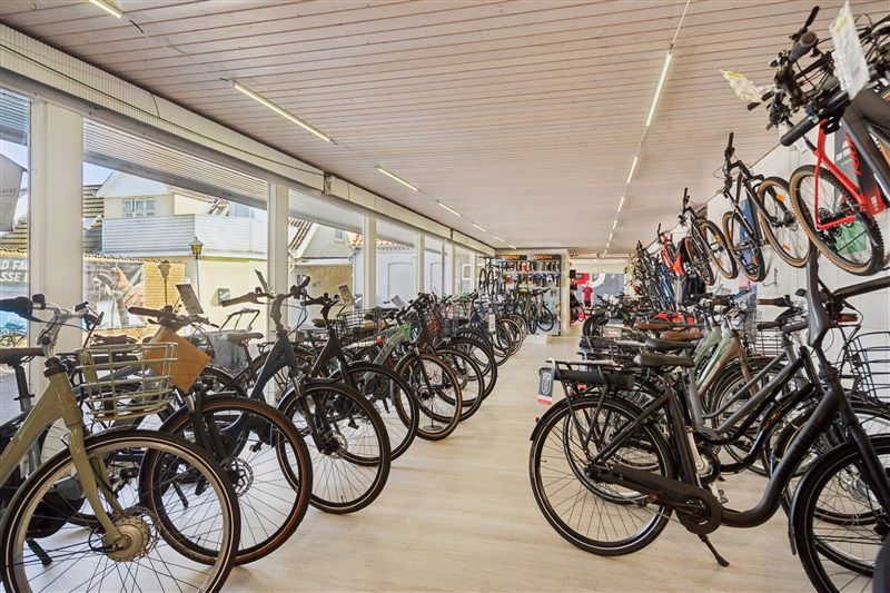 Mange cykler er parkeret på rækker i en forretning med lyse lofter og vinduer, der vender ud mod en bygning i baggrunden