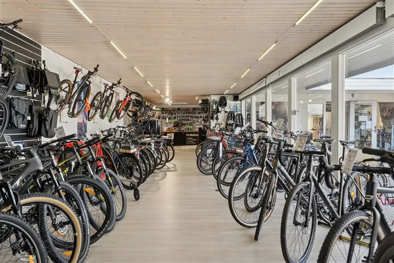 Mange cykler er udstillet i en cykelforretning med glasvægge, hængende cykler og mærkater.