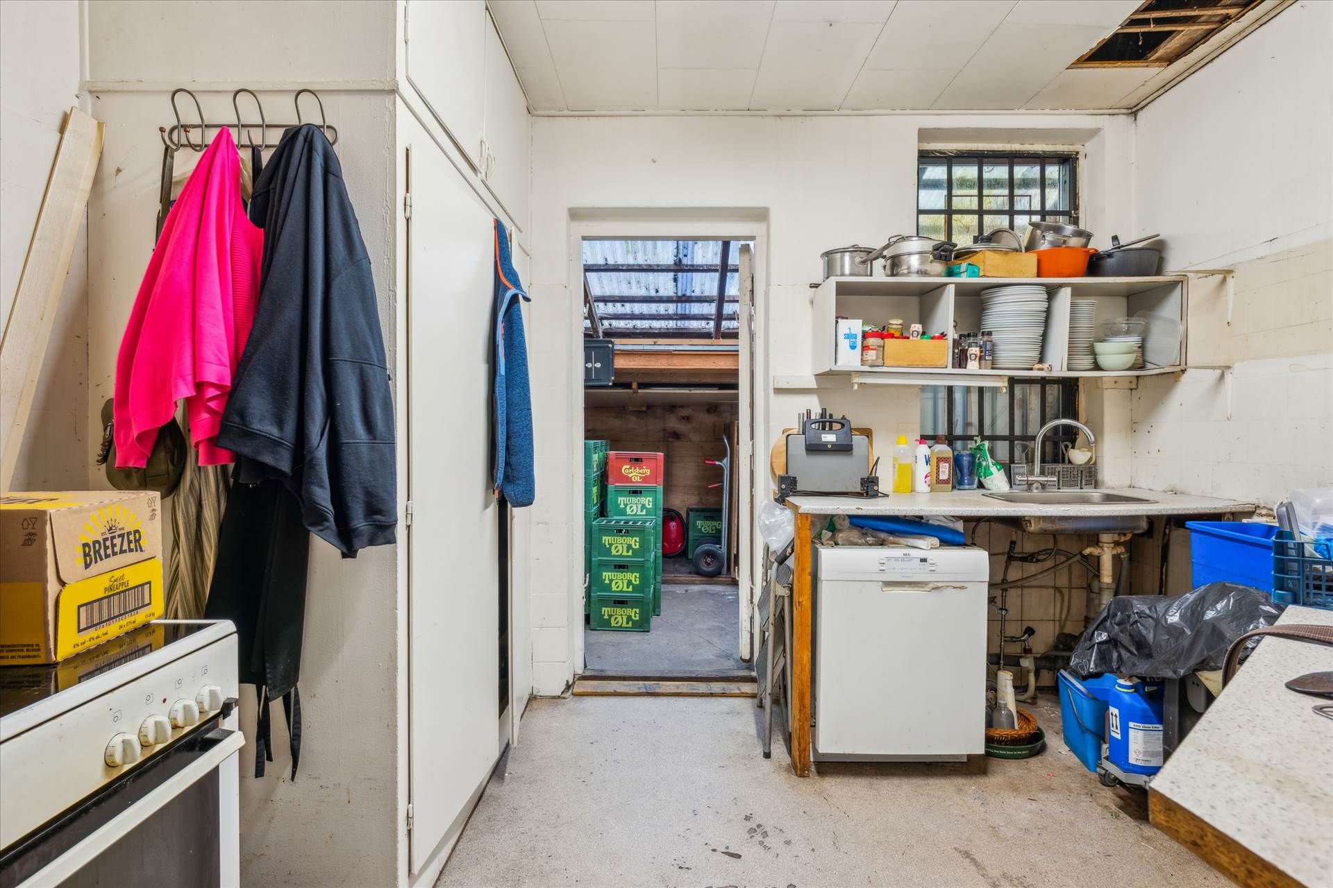 En ubenyttet køkkenrum med vaskemaskine, ovn og skrald. En åben dør fører til en lagerrum og en vindue.