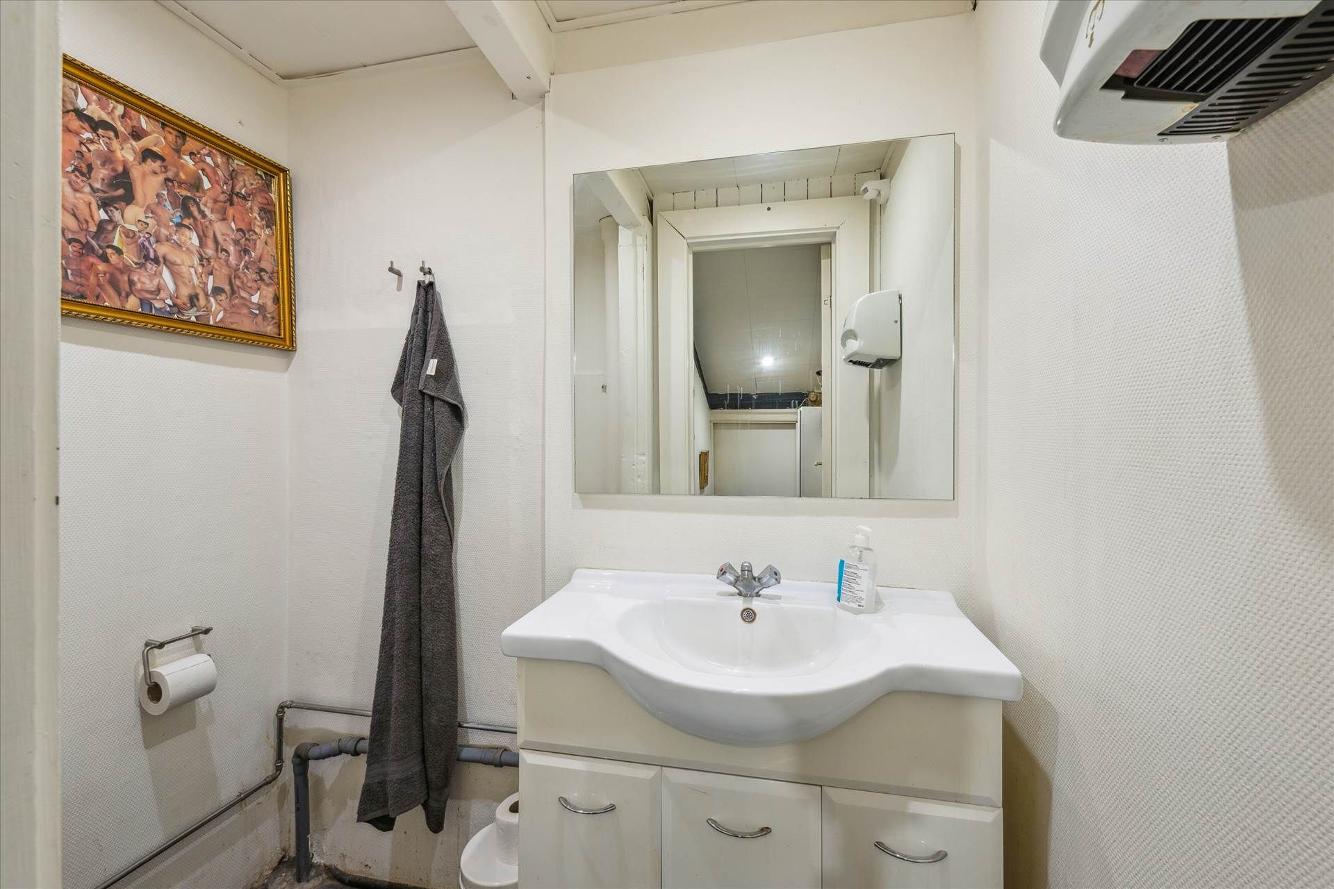 Et lille badeværelse med toilet, håndklæde, toiletpapir og en håndsæbe dispenser. Der er også et spejl og et klimaanlæg.