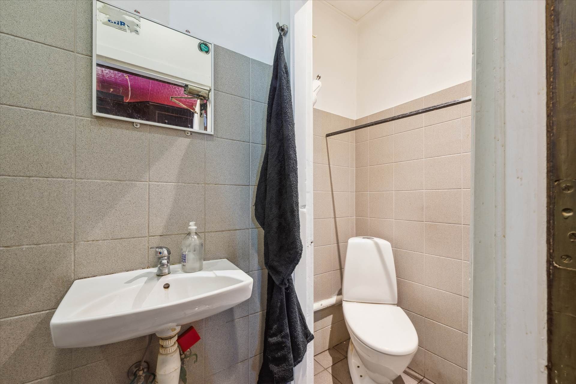 Et lille badeværelse med en hvid vask, en hvid toilet og en hængende sort håndklæde samt et spejl over vasken