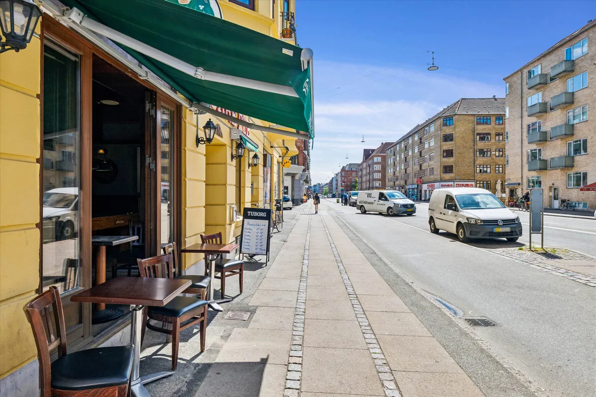 Butik med terrasse på Frederikssundsvej i Nordvestkvarter. To biler kører på vejen og to personer går på fortovet.