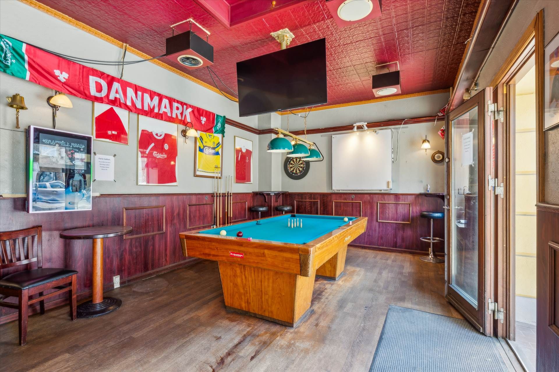 En poolbord og en dartplade er placeret i et lokale med røde vægge og et loft med pannelister og et TV-apparat