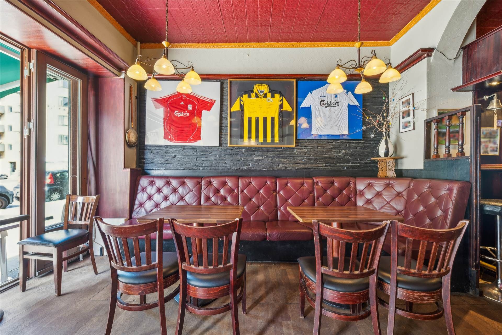 En restaurant med røde sofaer og træborde, hvor flere fodboldtrøyer er hængt op på væggen, og en lav bar i hjørnet med en
