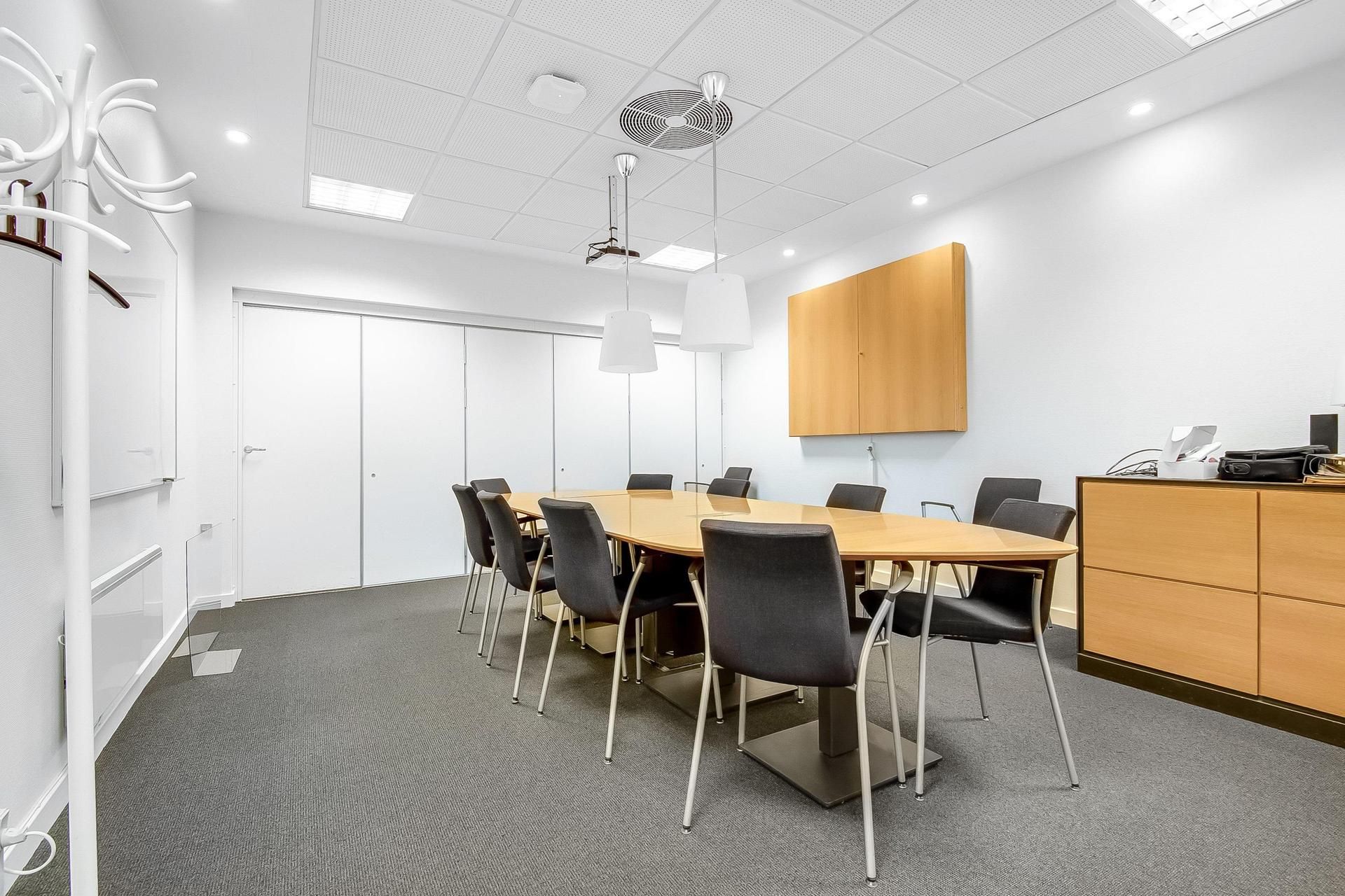 Her ses en mødelokale med lyse vægge og gulvtæppe, hvor der er en lang oval bord og stole, en projektor og en træskab med