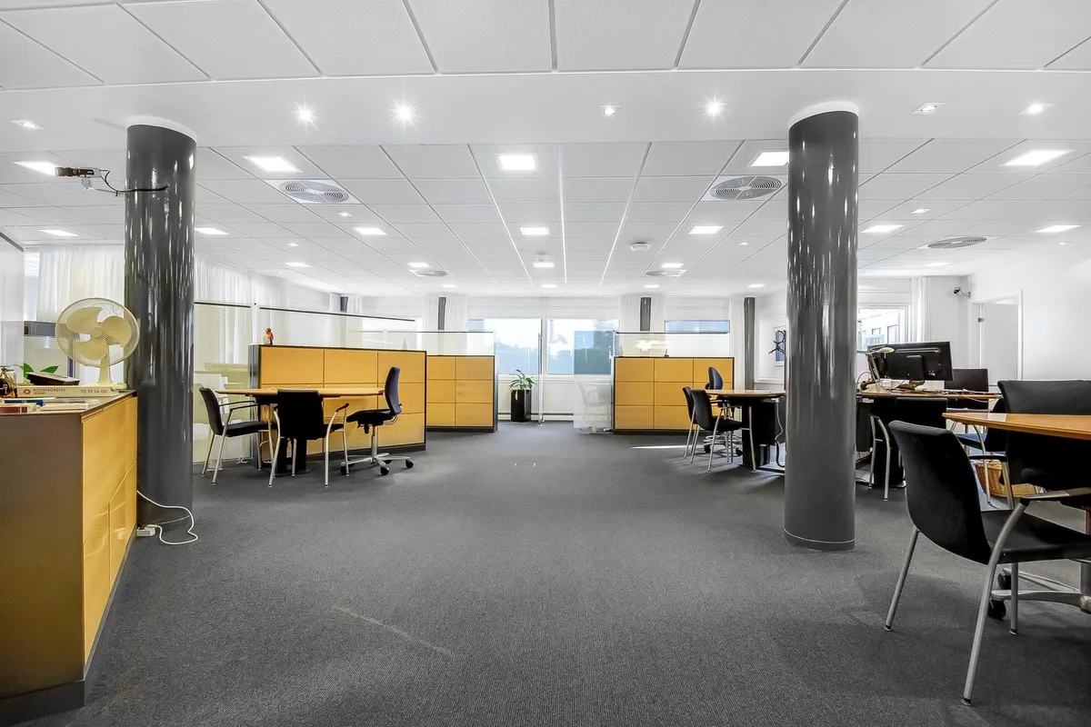 Et moderne kontor med lysegulve og hvide vægge, flere bordopsætninger med kontorstolar og computere, og gulbrune skillevægge