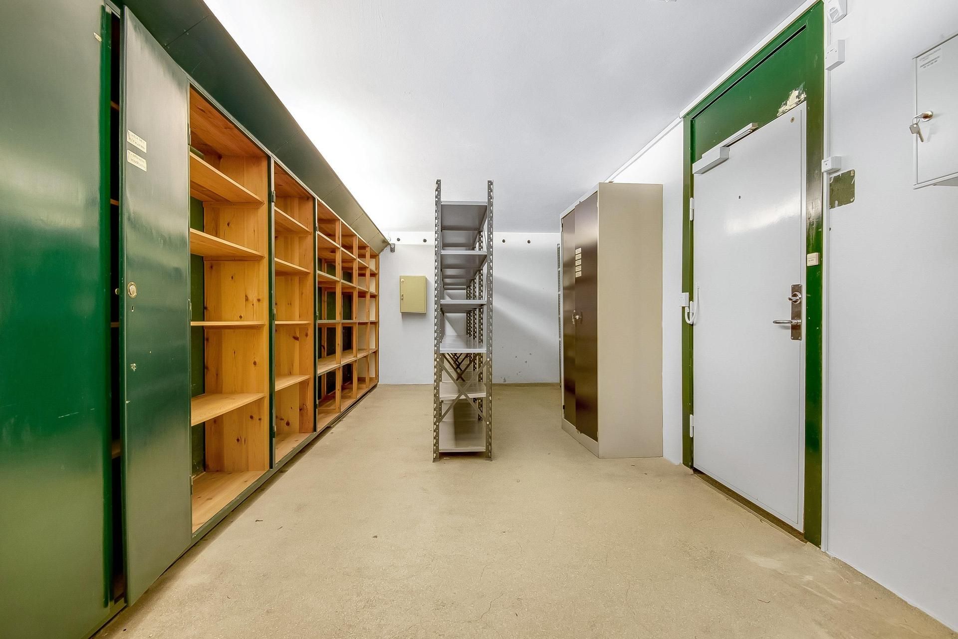 Lager med to rum og åbne grønne døre og hvide vægge og gulv, træreoler og metalhylder, samt en hvid dør med grøn indfatning