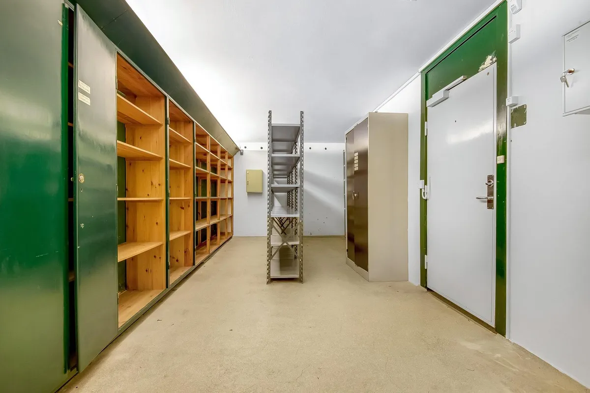 Lager med to rum og åbne grønne døre og hvide vægge og gulv, træreoler og metalhylder, samt en hvid dør med grøn indfatning