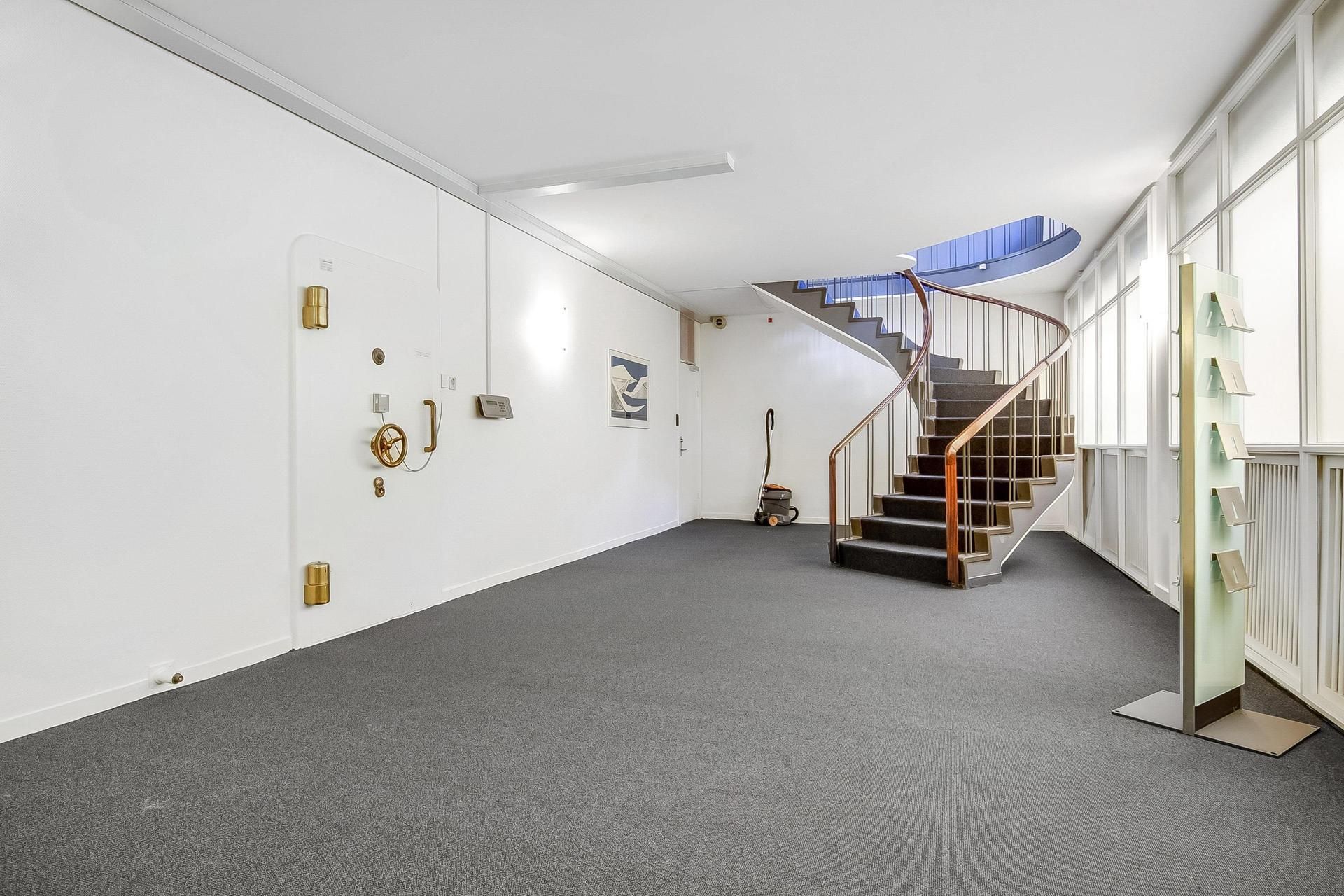 Et stort, tomt lokale med lysegrå gulvbelægning, spiraltrapper og hvide vægge samt en glasdør og et gulvmonteret display