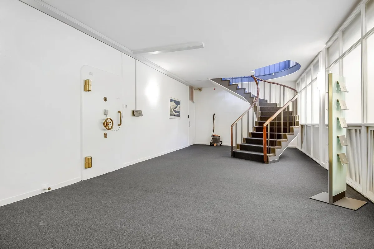 Et stort, tomt lokale med lysegrå gulvbelægning, spiraltrapper og hvide vægge samt en glasdør og et gulvmonteret display