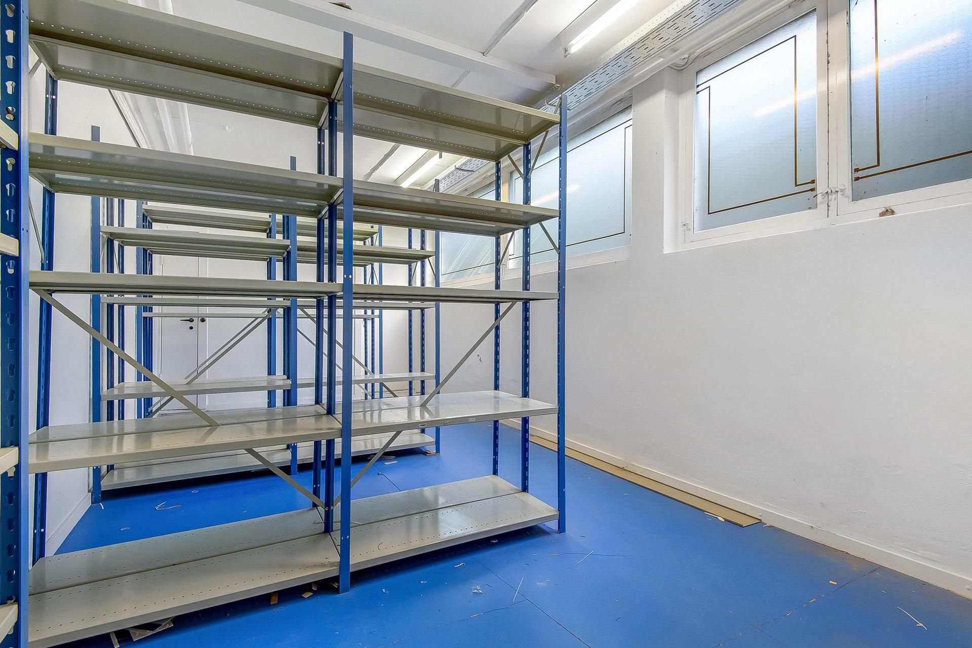 Lager med metalhylder i blå og hvid farve placeret i et hvidt lokale med vinduer og blå gulvbelægning