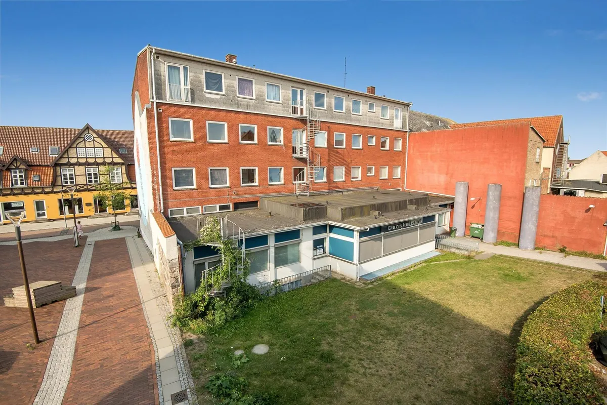 En bygning med hjørner af røde og hvide mursten og en indgangsdør, hvor en person går. Bagved er en lille have med græs og