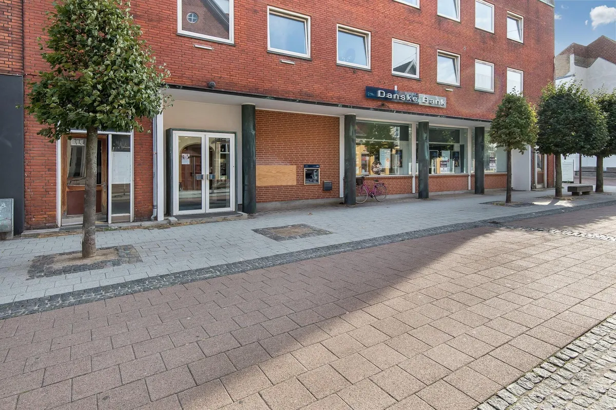 Foto af en forside med Danske Bank skilt, røde mursten og glasvinduer samt en lille plante og cykel