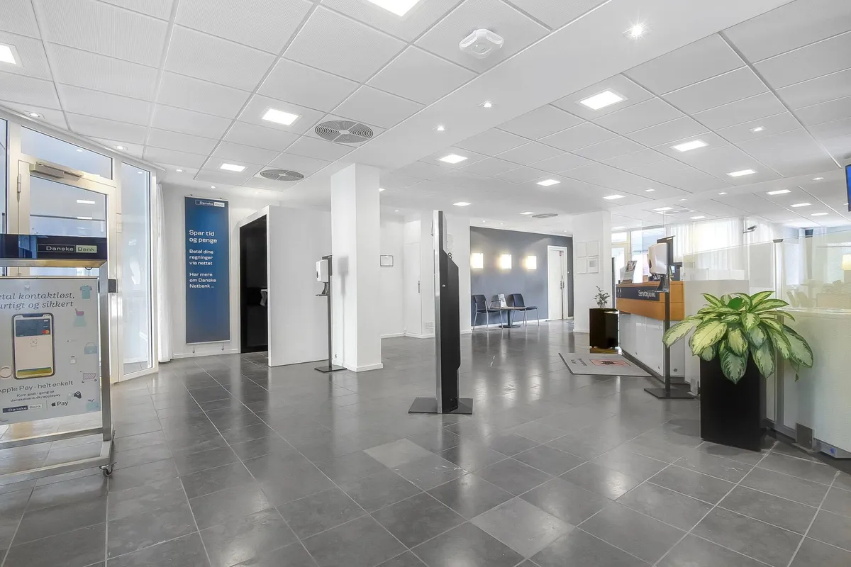 Store lokaler med højt loft, moderne møbler, gulv og vægge. To plantepoter med grønne planter og en stol til bagved et bord.