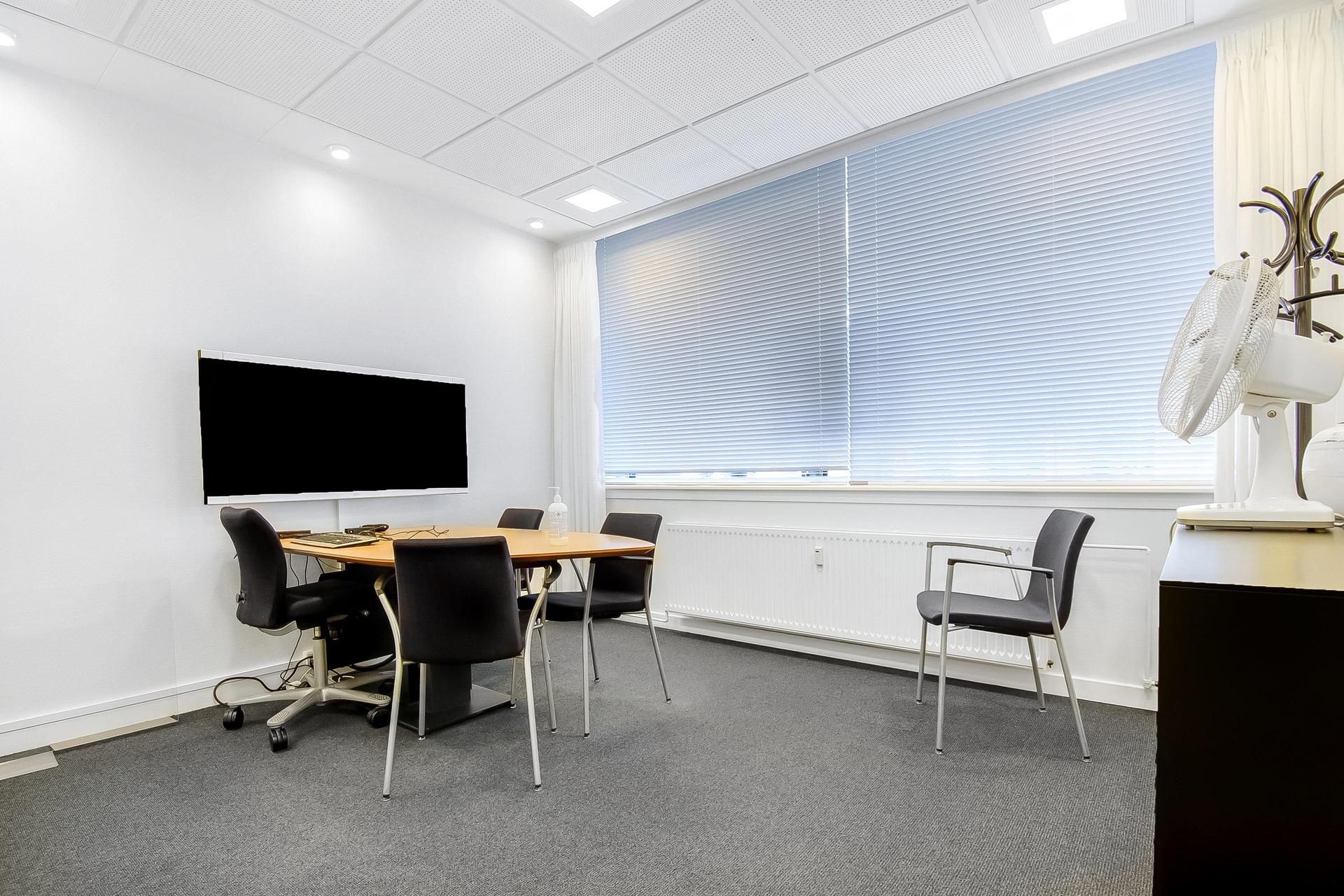 Mødelokalet har en stor skærm, bord og fire stole samt en ventilator ved vinduet