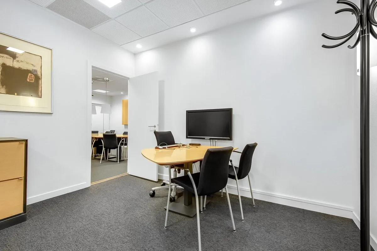 Det er et moderne mødelokale med lyse vægge, en fladt skærm og flere mødestoler med hjul. Der er en hængeklædeskab og et