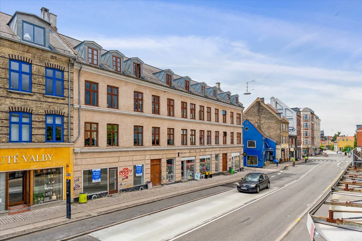 Erhvervslejlighed med butikslokaler på Valby Langgade. Forretninger, biler og folk vandrer rundt på gågaden og krydset.