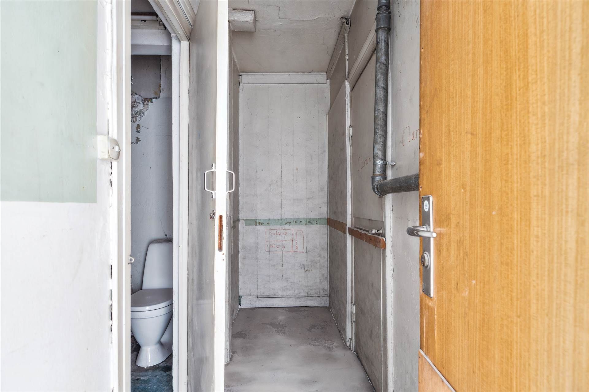 Et tomt badeværelse med åbne døre og en gammel toilet med skader på væggene og rør på væggen