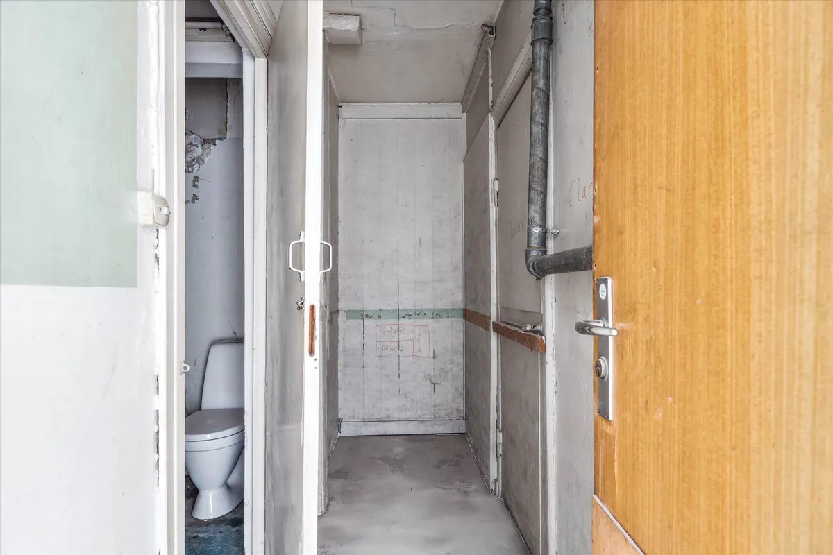 Et tomt badeværelse med åbne døre og en gammel toilet med skader på væggene og rør på væggen