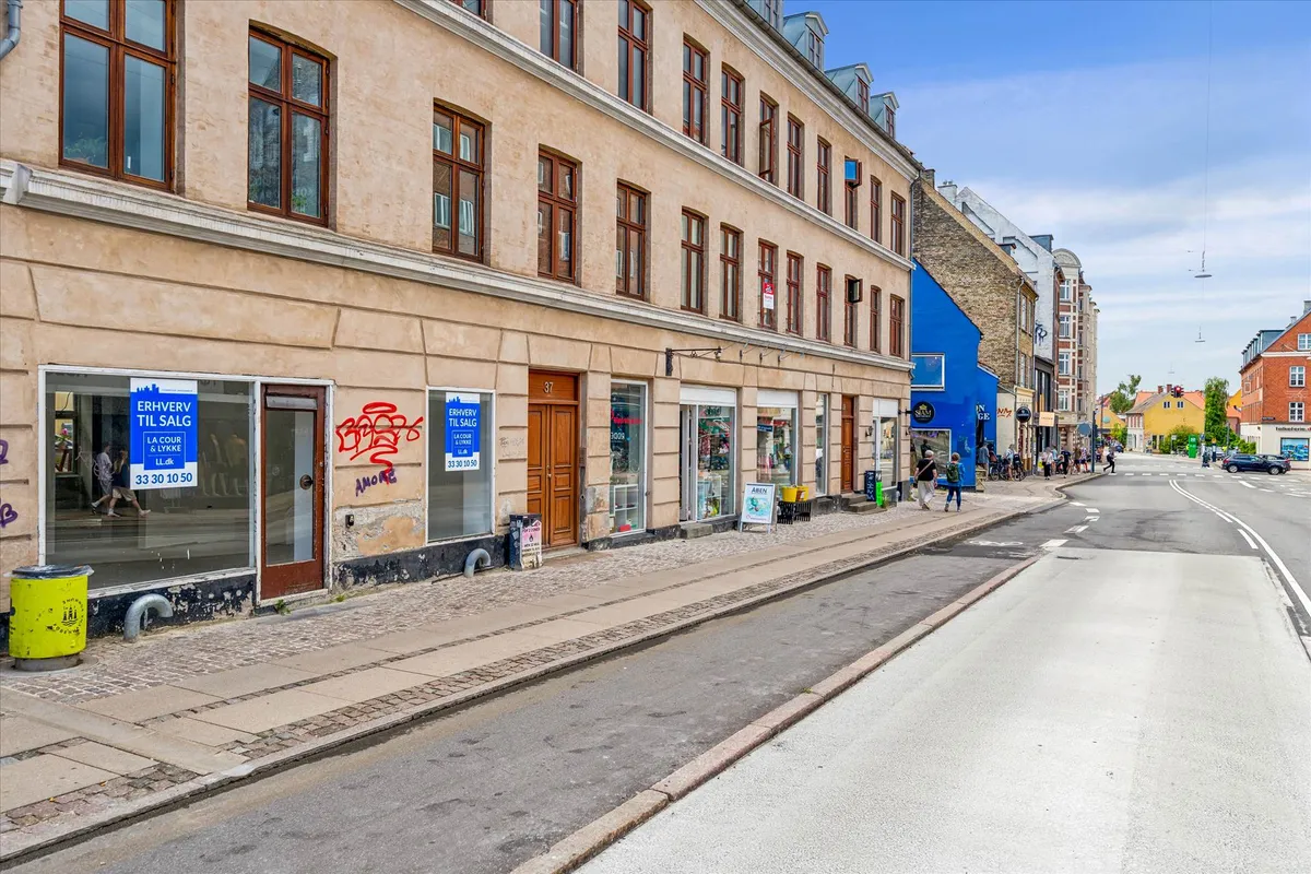 Erhvervsejlejlighed med oplysning om salg, glasvinduer, skraldespand og graffiti på en bygning.