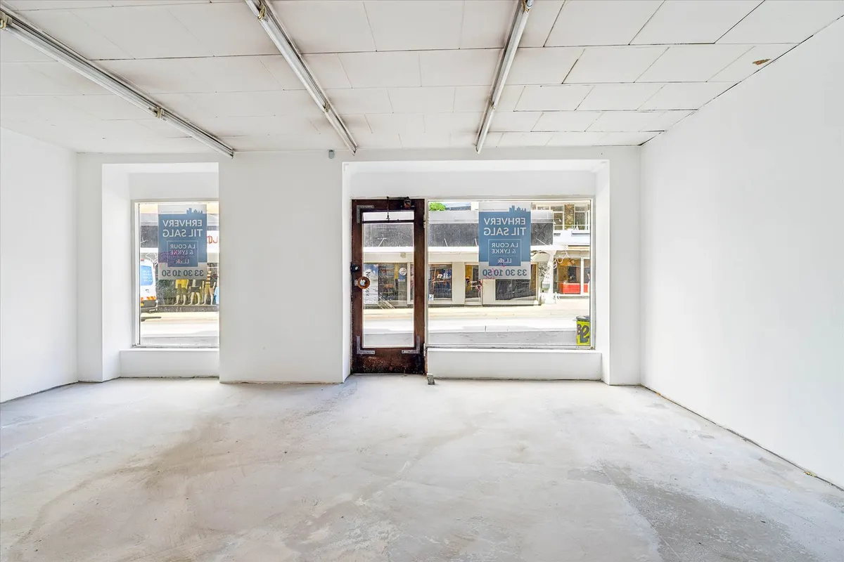 Indvendigt i en tom erhvervslejlighed med hvide vægge og lyse hvide lofter med lysebuelys. To vinduer og en dør med