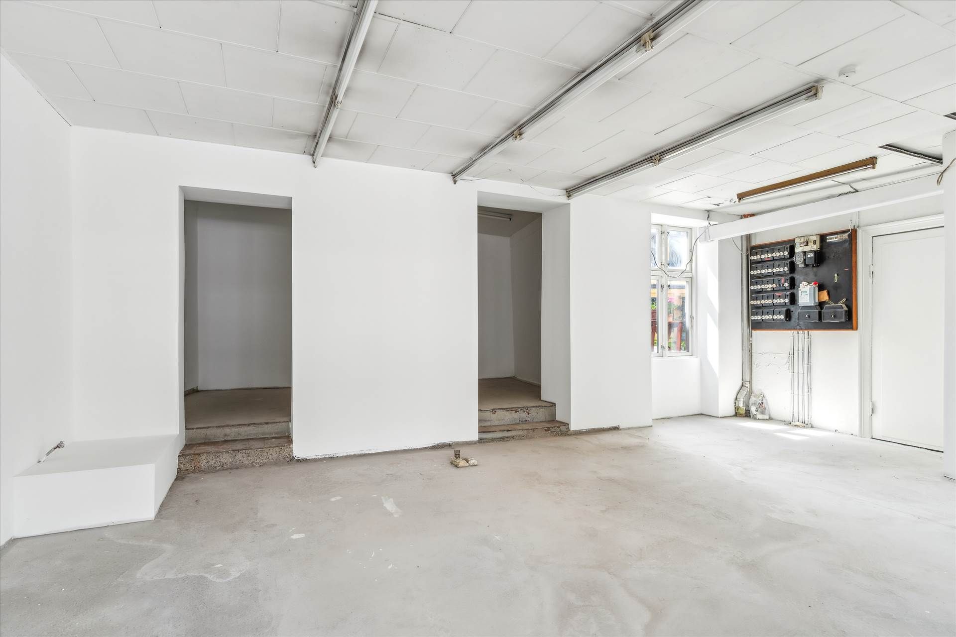 Store, hvidmalet lokale med vinduer, udvendigt elektriske kontakter og en indgangsdør. Lokaler med betongulv og to
