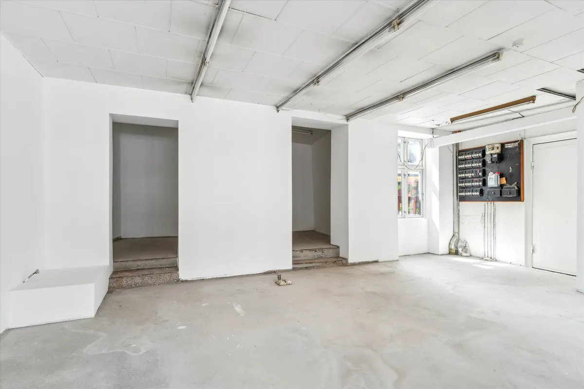 Store, hvidmalet lokale med vinduer, udvendigt elektriske kontakter og en indgangsdør. Lokaler med betongulv og to