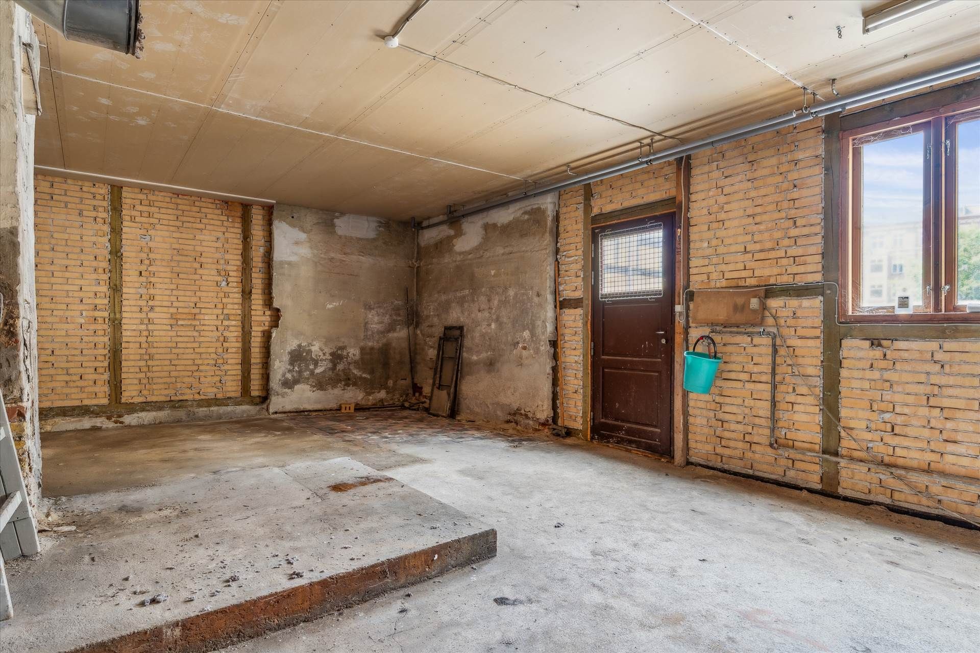 Forladt garage med gamle mursten og betongulv, lyseblå spand hængende på væggen, udsigt til gaden gennem vinduet og trækasse