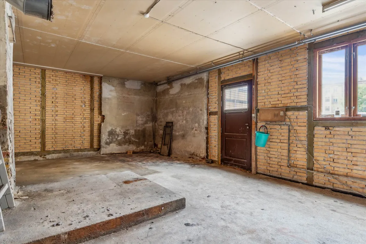 Forladt garage med gamle mursten og betongulv, lyseblå spand hængende på væggen, udsigt til gaden gennem vinduet og trækasse