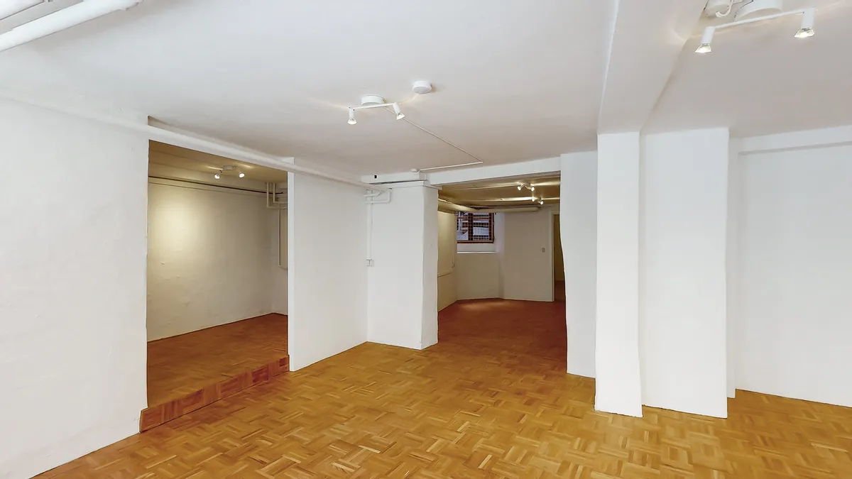 Værelse med hvide vægge og lyse gulv. Lysekroner er monteret på loftet. Et hul fører til et andet rum med gulv og højt