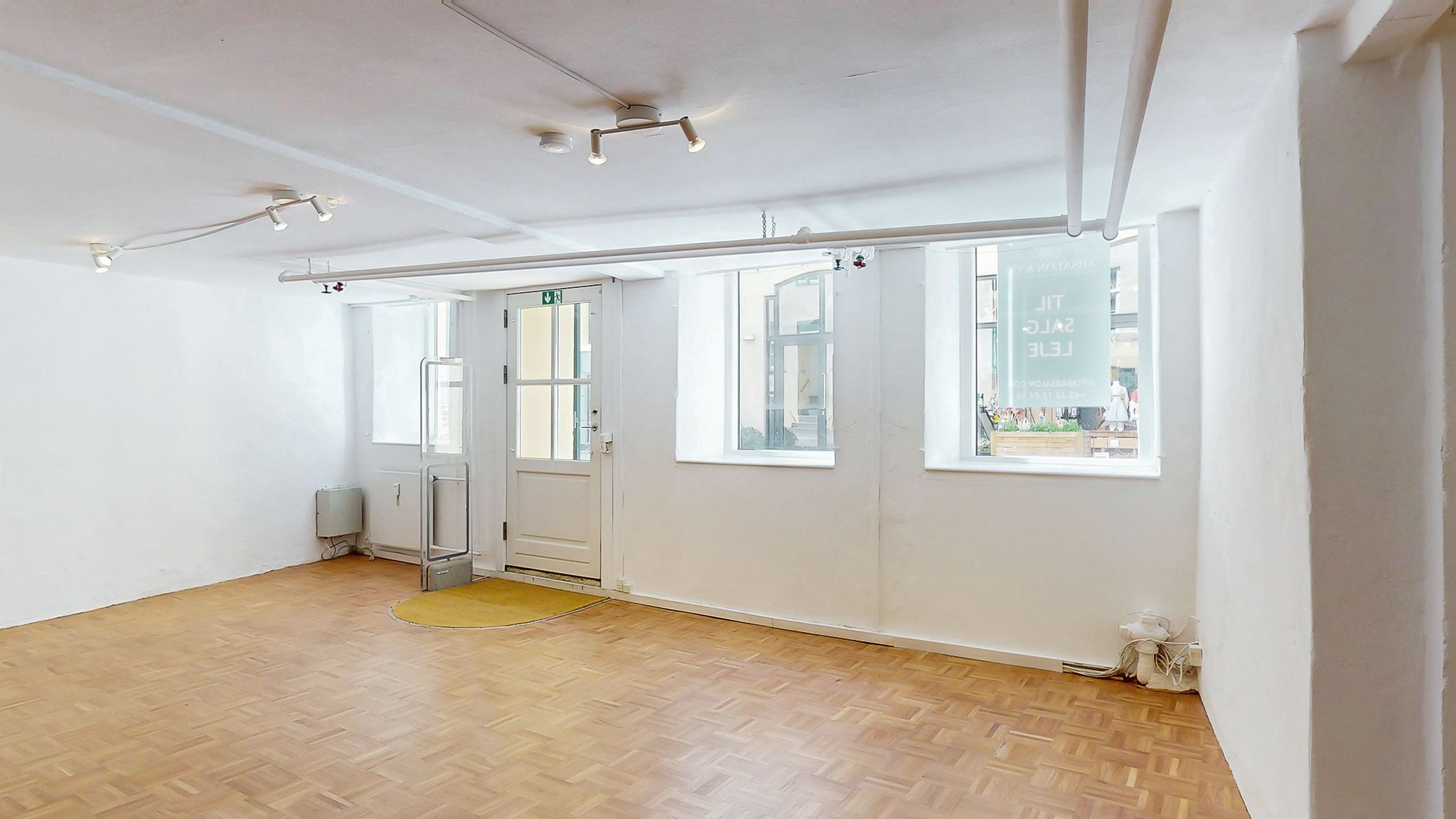 Tomt lokale med lysegul gulv og hvide vægge. Der er tre vinduer med samme udsigt til gaden og et banner med teksten Let til