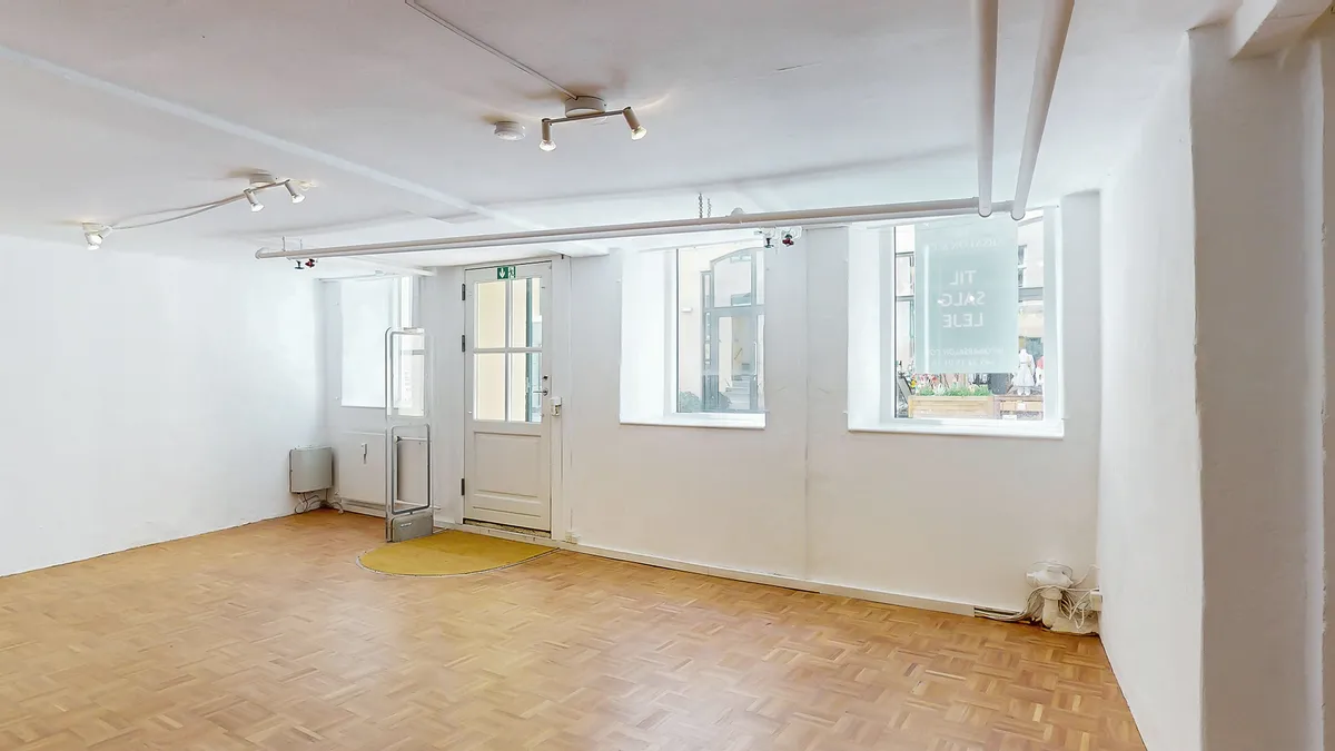 Tomt lokale med lysegul gulv og hvide vægge. Der er tre vinduer med samme udsigt til gaden og et banner med teksten Let til