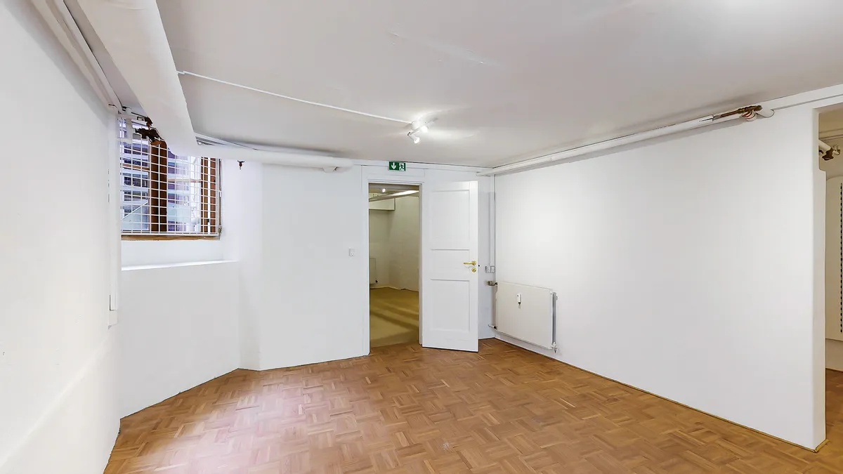 En tom rum med hvide vægge og trægulv samt åbne døre og et vindue med gitter i baggrunden